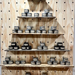 May include: A display of numerous small, round metal candle tins with labels, arranged on five horizontal wooden shelves attached to a large light-colored pegboard. A decorative star-shaped ornament with pinecones sits on the top shelf.