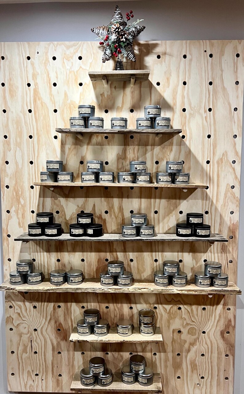 May include: A display of numerous small, round metal candle tins with labels, arranged on five horizontal wooden shelves attached to a large light-colored pegboard. A decorative star-shaped ornament with pinecones sits on the top shelf.