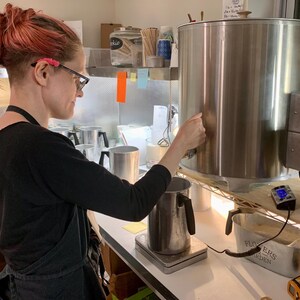 May include: A large stainless steel wax melting tank with a spigot and temperature control dials, positioned next to a metal pouring pitcher on a digital scale, with a small metal container labeled Flowers and Garden nearby on the counter.
