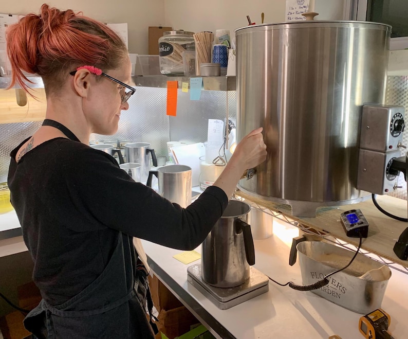 May include: A large stainless steel wax melting tank with a spigot and temperature control dials, positioned next to a metal pouring pitcher on a digital scale, with a small metal container labeled Flowers and Garden nearby on the counter.