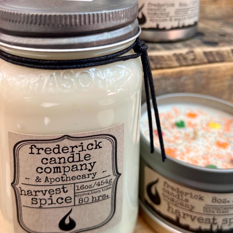 May include: A 16oz glass mason jar candle with a silver metal lid and a black string tied around the neck. The label reads Frederick Candle Company & Apothecary, Harvest Spice, 16oz/454g, 80 hrs. A smaller 8oz metal tin candle is visible nearby.