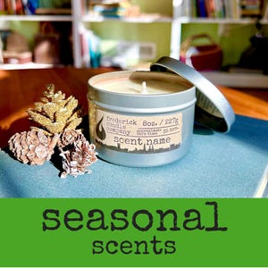 May include: A silver metal candle tin with a lid and a label reading frederick candle company 8oz. / 227g approximate burn time: 35 hrs. scent name, accompanied by three gold-toned pinecones, resting on a blue surface with text below.