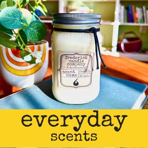 May include: A white candle in a clear glass jar with a silver metal lid and a black string tied around the neck. The label reads Frederick Candle Company & Apothecary, Scent Name, 16oz/454g, and 80 hrs. The candle sits on a blue book.