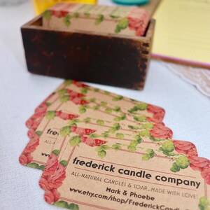 May include: A stack of rectangular business cards with rounded corners featuring a floral border of red roses and green leaves on kraft paper. The text reads frederick candle company, ALL-NATURAL CANDLES & SOAP MADE WITH LOVE, Mark & Phoebe.