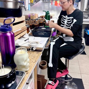 May include: A black long-sleeve shirt and matching pants featuring a white skeleton bone print. The outfit is worn with bright pink slide sandals and patterned socks. The shirt has a crew neckline and the pants have a straight-leg fit.