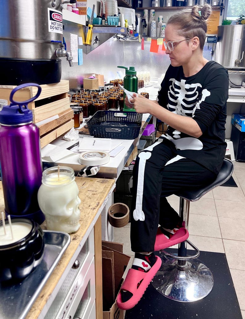 May include: A black long-sleeve shirt and matching pants featuring a white skeleton bone print. The outfit is worn with bright pink slide sandals and patterned socks. The shirt has a crew neckline and the pants have a straight-leg fit.
