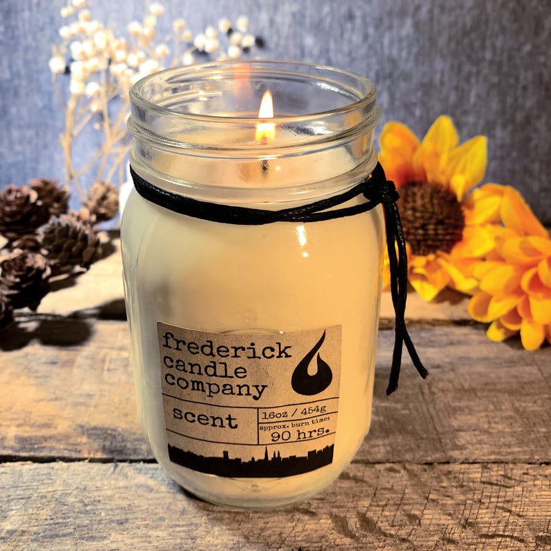 May include: A lit candle in a clear glass mason jar with a black string tied around the rim. The label reads frederick candle company, scent, 16oz / 454g, and approx. burn time 90 hrs. The wax is off-white and the jar has a visible flame.