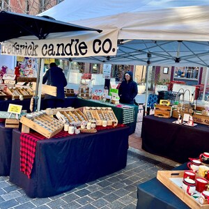 May include: An outdoor market stall with a white canopy featuring the text Frederick Candle Co. The stall displays various candles in glass jars arranged in wooden crates on tables covered with black cloths and red and black plaid runners.