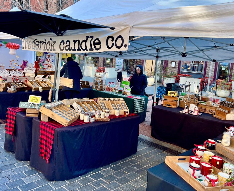 May include: An outdoor market stall with a white canopy featuring the text Frederick Candle Co. The stall displays various candles in glass jars arranged in wooden crates on tables covered with black cloths and red and black plaid runners.