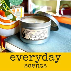 May include: A silver metal candle tin with a white wax interior and a single wick. The label reads: frederick candle company, 8oz. / 227g, approximate burn time: 35 hrs., scent name. The lid is partially open, resting beside the candle container.