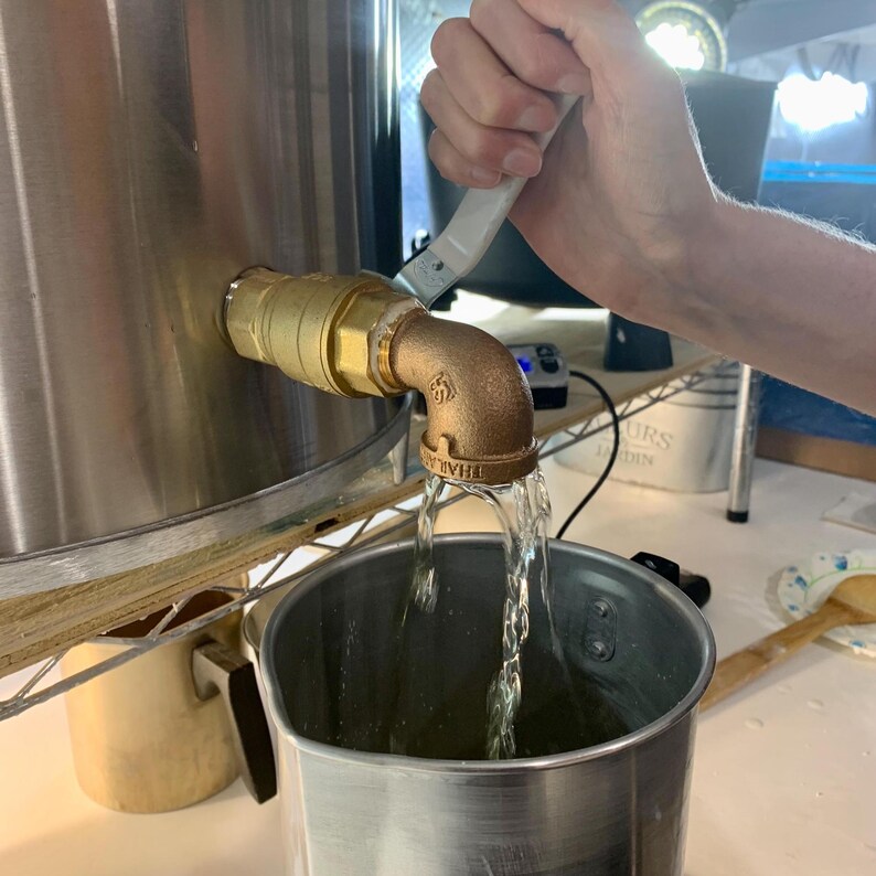 May include: A stainless steel vessel with a brass ball valve spigot pouring a clear liquid into a stainless steel pot. The spigot features a white handle and a threaded elbow fitting, with the liquid flowing in a steady stream from the spout.
