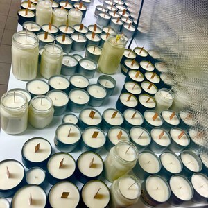 May include: A large collection of candles in various containers, including clear glass jars and small metal tins. Each candle features a smooth, off-white wax surface with a centered wick, some being wooden and others being traditional string wicks.