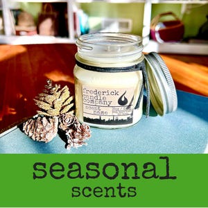 May include: A glass jar candle with a cream-colored wax and a black string tied around the neck. The label reads Frederick Candle Company, Scent Name, 8oz / 227g, and 50 hrs. burn time. Three small pinecones sit beside the jar, which has a silver lid.