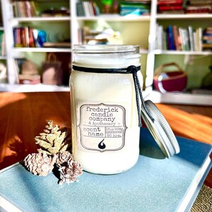 May include: A white candle in a clear glass mason jar with a silver lid and a black string tied around the neck. The label reads Frederick Candle Company & Apothecary, 16oz/454g, 80 hrs. Three small, light brown pinecones sit next to the jar.