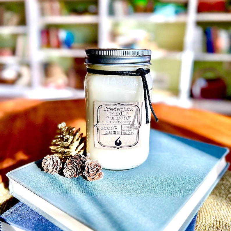 May include: A white candle in a clear glass jar with a silver metal lid and a black string tied around the neck. The label reads Frederick Candle Company & Apothecary, Scent Name, 16oz/454g, 90 hrs. Three small pinecones are placed at the base.