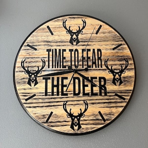 May include: A wooden clock with a black face and black hands. The clock has a wood grain pattern and the words "Time to Fear the Deer" are printed on the face. There are four black deer silhouettes on the clock face.