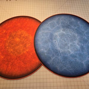 Peut inclure: Deux plateaux de jeu ronds et plats. Un plateau est orange et rouge avec une bordure brun fonc&eacute;. L'autre plateau est bleu avec un motif marbr&eacute; blanc et une bordure brun fonc&eacute;.