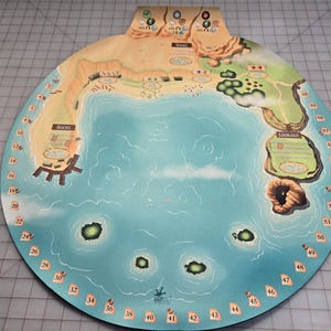 May include: A circular board game map with a coastal theme. The map features a blue ocean, islands, and landmasses with docks and lookout points. Numbers are printed around the edge of the board.