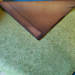 May include: A green felt gaming mat with a brown felt top folded over it. The mat is folded in half and the brown felt is on top.