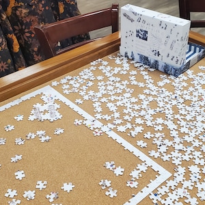 May include: A partially completed jigsaw puzzle on a wooden table. The puzzle pieces are white with a variety of shapes. A box with a blue and white design sits nearby. The puzzle is on a corkboard surface.
