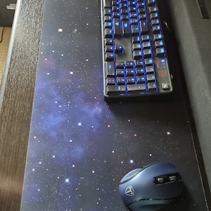 May include: A black and blue gaming keyboard with blue backlit keys rests on a large mousepad. The mousepad features a starry night design with a blue wireless mouse. The keyboard and mouse are on a dark surface.
