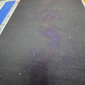 Puede incluir: Una tela gris oscura con un diseño de galaxia que presenta una nebulosa púrpura y rosa con estrellas blancas y amarillas.