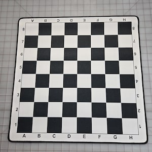 May include: A black and white checkered chess board with letters and numbers along the edges. The board is rectangular with a black border and is set against a gray background.