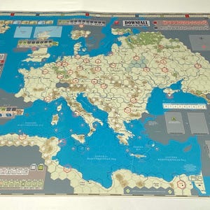 May include: A board game map depicting Europe and surrounding regions, with a blue sea and landmasses in beige and green. The game board includes text such as "DOWNFALL" and various game elements.