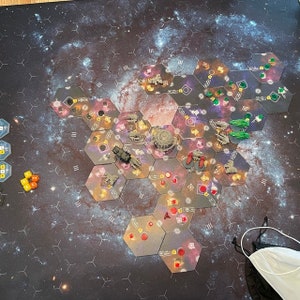 May include: A galaxy-themed game board with hexagonal tiles, featuring spaceships, dice, and other game pieces. The board is set against a dark background with a swirling galaxy pattern.