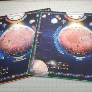 May include: Two game boards for the board game "Terraforming Mars". The boards are black with a space theme and feature a red planet with a hexagonal grid. One board has an orange border and the other has a green border.