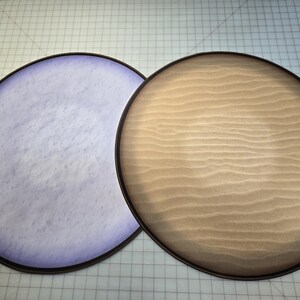 Peut inclure: Deux plateaux ronds en bois plats avec un bord noir. Un plateau a une surface marbr&eacute;e violet clair et l'autre a une surface en bois brun clair.