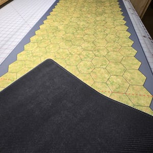 Puede incluir: Una alfombrilla de juego rectangular grande con un patrón de cuadrícula hexagonal. La alfombrilla presenta un diseño de paisaje verde y amarillo con un borde gris. La parte inferior de la alfombrilla es negra. Diseñada para juegos de mesa.