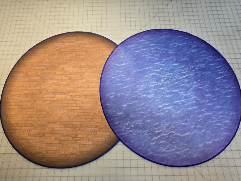 Peut inclure: Deux tapis de jeu ronds, &agrave; dos en feutre. Un tapis est brun avec un motif en bois, l'autre est bleu avec un motif de vagues.