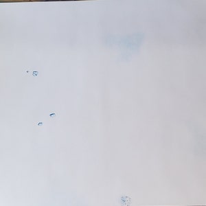 May include: A white sheet of paper with blue paint splatters. The splatters are of various sizes and shapes. The paper is slightly creased.