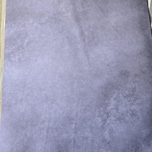May include: A sheet of textured paper in shades of lavender and grey. The paper has a rough, uneven surface with darker areas creating a subtle, aged appearance. The overall effect is a muted, vintage aesthetic.