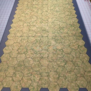 May include: A rectangular gaming mat featuring a printed grid of tan and green hexagonal tiles. The mat has a solid dark grey border surrounding the central hexagonal pattern. Two horizontal dashed lines cross the grid at regular intervals.