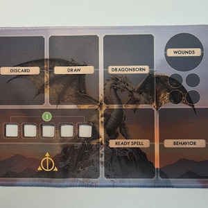 Puede incluir: Un tapete de juego rectangular con una ilustración central de un dragón sobre un paisaje montañoso. El tapete incluye secciones etiquetadas DISCARD, DRAW, DRAGONBORN, WOUNDS, READY SPELL y BEHAVIOR, junto a cinco cuadrados blancos.