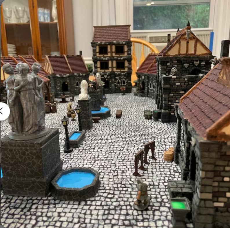 May include: A miniature medieval town square with stone buildings, a fountain, and a cobbled street. The buildings have red tile roofs and are decorated with stone details. There are small figures of people and animals throughout the scene.