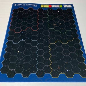 Puede incluir: Una alfombrilla de juego rectangular azul con un diseño de cuadrícula hexagonal. La alfombrilla presenta el texto "SPACE EMPIRES" en la parte superior y una serie de cuadros de colores. La cuadrícula es negra con líneas de puntos blancas y un tema espacial.