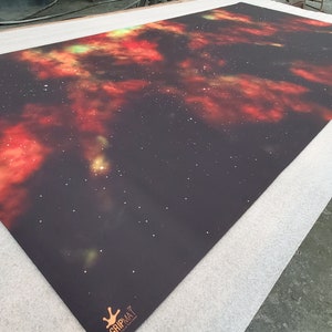 Puede incluir: Una alfombrilla de juego negra con un diseño de nebulosa en colores rojo, naranja y verde. La alfombrilla tiene un efecto de brillo sutil y el texto "GripMat" en la esquina inferior derecha.