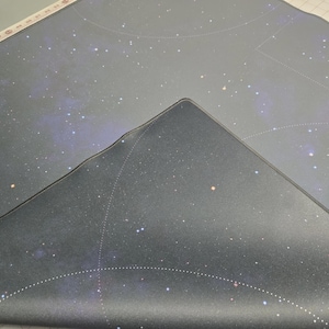 May include: A dark gray, rectangular mat with a celestial design featuring white stars and constellations. The mat is partially folded, revealing a smooth, dark surface with a subtle sheen. The design evokes a sense of the cosmos.