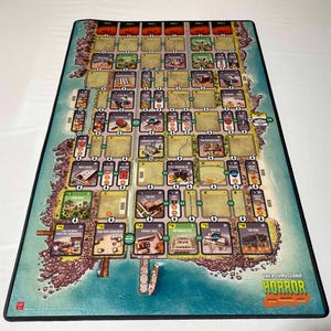 Puede incluir: Un tapete de juego rectangular con un mapa de una isla, con varias ubicaciones y elementos del juego. El tapete tiene un borde turquesa y el título "THE PLUM ISLAND HORROR" en texto naranja y verde.