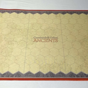 Puede incluir: Un tablero de juego rectangular con un patrón de cuadrícula hexagonal. El tablero tiene una superficie beige con un borde rojo. El texto "Commands & Colors ANCIENTS" está impreso en el centro del tablero.