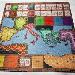 Op de afbeelding: Een bordspel met een kaart van een regio, met zeshoekige gebieden in verschillende kleuren. Het spelbord heeft kaarten bovenaan en tekstlabels onderaan. Het bord is omgeven door een baksteenachtige rand.