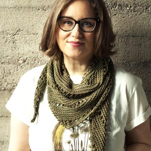 May include: A woman wearing a white t-shirt with a botanical print and a green knitted shawl. The shawl is draped around the neck and shoulders. She is wearing black-framed glasses.