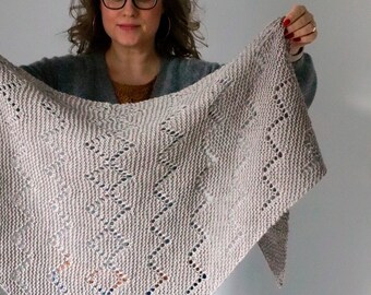May include: A woman is holding up a light gray knitted shawl with a lacy pattern. The shawl is triangular in shape and has a delicate, openwork design.