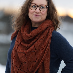 May include: A rust-colored triangular knit shawl featuring a geometric lace pattern with eyelet details. The shawl is draped over a dark navy blue long-sleeved sweater. The knit fabric has a soft texture and is wrapped around the neck and shoulders.
