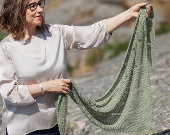 Op de afbeelding: Een vrouw in een lichtbeige top en blauwe jeans houdt een groene gebreide sjaal met een kantpatroon vast.