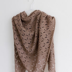 May include: A brown knitted shawl with a lacy pattern hangs on a white hanger against a white background.