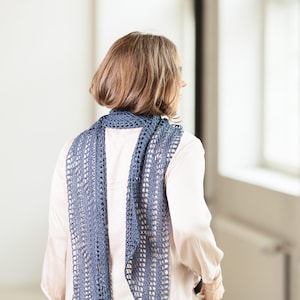 Peut inclure: Une écharpe bleue crochetée, avec un motif ajouré, est portée sur les épaules. La personne porte un chemisier rose clair et un jean bleu. L'arrière-plan est un espace clair et neutre. C'est un accessoire de mode.
